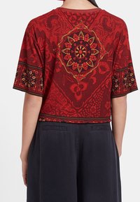 Röd mönstrad kortärmad topp med blommönster och guldaccenter, med rundad halsringning och avslappnad passform. Matchad med mörka byxor.