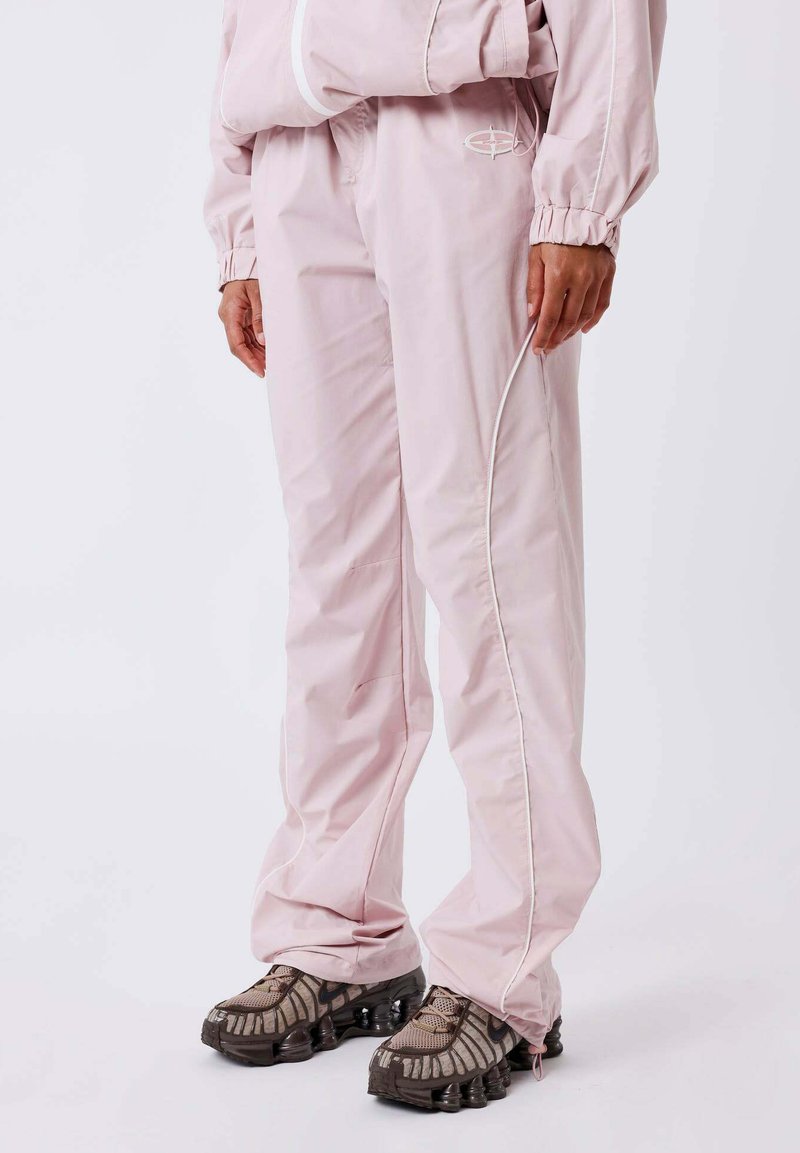Persona vestida con un chándal rosa claro, pantalones y chaqueta, de pie, con zapatillas deportivas marrón y negro sobre un fondo blanco.