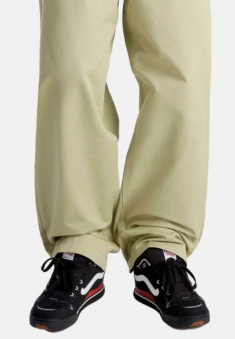 Zwarte sneakers met witte en rode accenten, en beige wijde broek. De sneakers hebben een gestructureerde zwarte bovenzijde en vetersluiting.