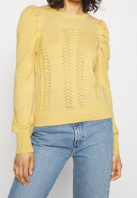 Pull en maille jaune à manches bouffantes, avec un motif texturé et des poignets et un ourlet côtelés, associé à un jean bleu clair.