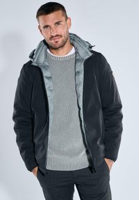 Black jacket with a gray puffer interior, zippered front, and adjustable hood, worn over a light gray textured knit sweater.