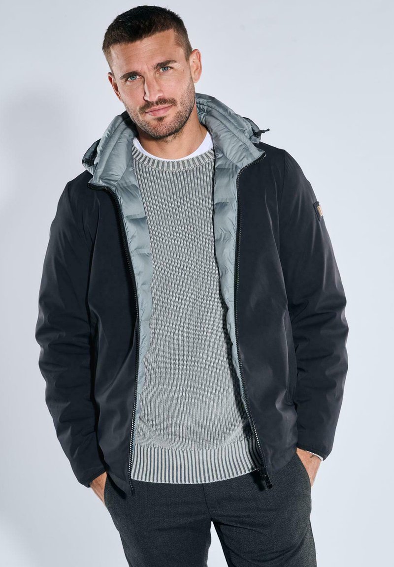 Black jacket with a gray puffer interior, zippered front, and adjustable hood, worn over a light gray textured knit sweater.