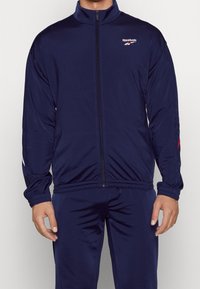 Tmavě modrá tepláková bunda z hladké látky, s vysokým límcem, celým zipem a logem Reebok. Výrazné červené a bílé akcenty na ramenech.