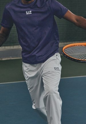 Homme en chemise bleue et pantalon blanc jouant au tennis, tenant une raquette avec un bord orange sur un court intérieur.