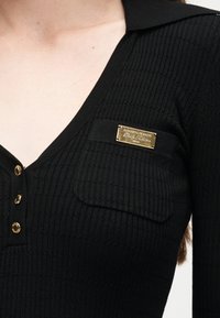 Κοντινό πλάνο μαύρου πλεκτού τοπ με ριμπ ύφανση, με χρυσά κουμπιά μπροστά και χρυσή πλακέτα με το λογότυπο της μάρκας στην τσέπη στο στήθος.