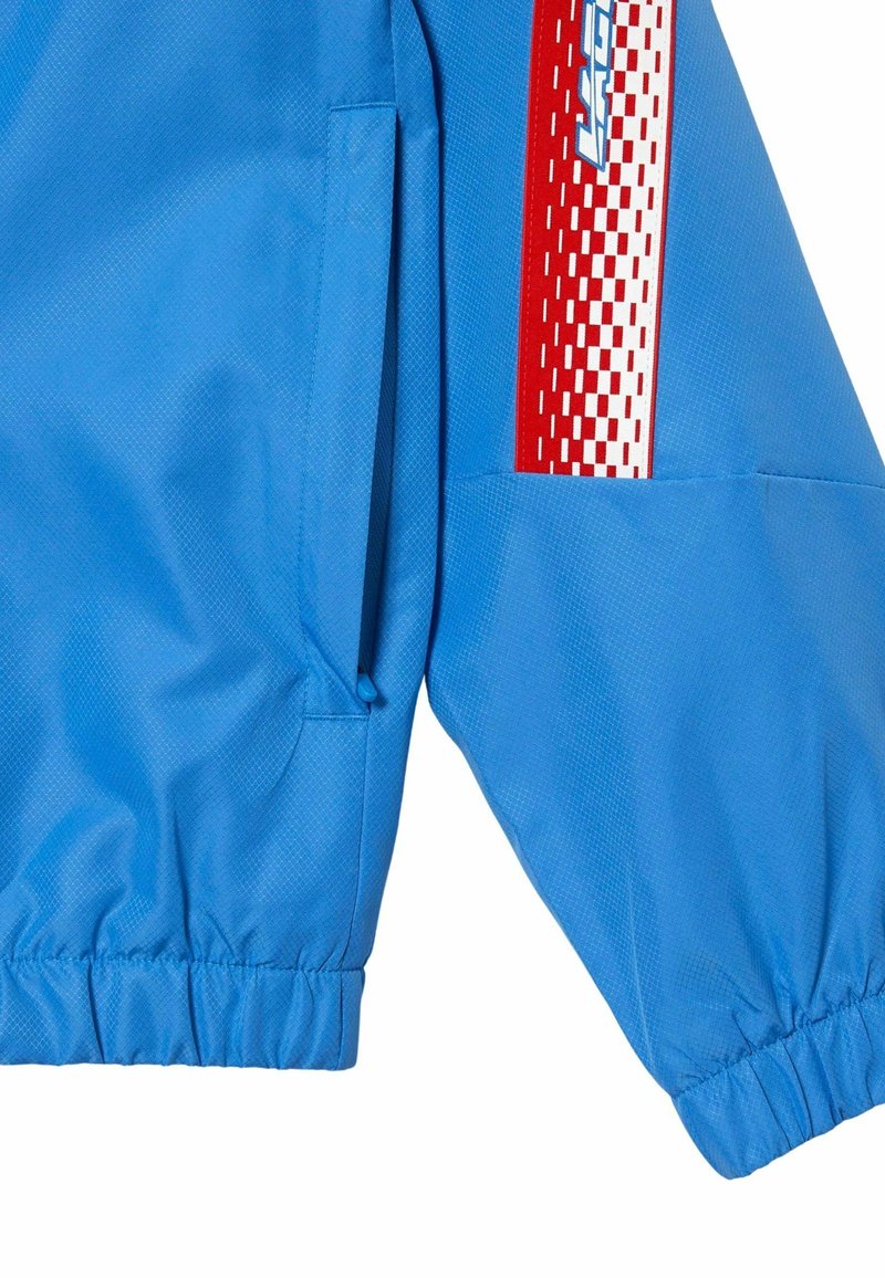 Veste légère bleue avec un tissu texturé, présentant un accent à carreaux rouge et blanc le long de la manche et des poignets élastiqués pour un ajustement parfait.