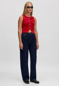 Rotes ärmelloses Oberteil mit Knopfdetails, kombiniert mit dunkelblauen, gerade geschnittenen Jeans. Die Accessoires umfassen schwarze ovale Sonnenbrillen. Glatte Texturen.