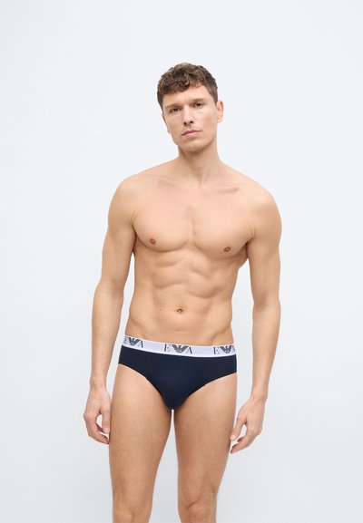 Särgi seljast võtnud mees, lühikeste lokkis juustega, kandes tumesiniseid pükse valge EA logoga vöökohal, seisab ühevärvilise heleda tausta ees.