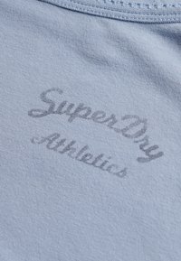 Tela azul claro con texto gris oscuro desvanecido que dice "SuperDry Athletics" en una fuente cursiva cerca de una costura del escote.