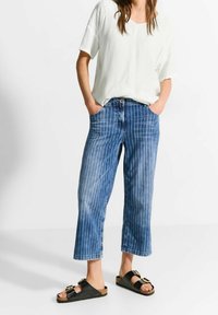 Vit kortärmad topp kombinerad med blårandiga vida jeans och svarta sandaler med spänne. Har framliga fickor och en avslappnad passform.