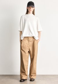 Przewymiarowany biały bawełniany t-shirt, beżowe bawełniane spodnie o szerokich nogawkach, czapka w kolorze czarnym oraz brązowe skórzane buty. Prosty design, casualowe dopasowanie.