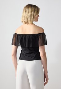 Top negru, cu umerii goi, cu un strat transparent și volanat, siluetă ajustată și textură netedă. Purtați cu pantaloni albi, cu talie înaltă.