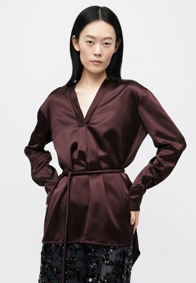 Γυναίκα με ίσια μαύρα μαλλιά φοράει βαθιά βυσσινί σατινέ μπλούζα με ζώνη, συνδυασμένη με μαύρη φούστα με παγιέτες, στέκεται με τα χέρια στη μέση.