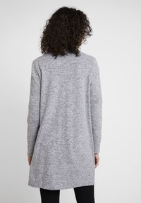 Gilet en tricot gris avec une coupe mi-longue, des manches longues et une coupe décontractée. Le tissu texturé accentue la douceur générale. Pas de fermetures visibles.