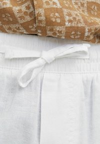 Un tissu marron à motifs tombe sur une taille en lin blanc avec un nœud noué, présentant une finition texturée et un détail plissé en dessous.