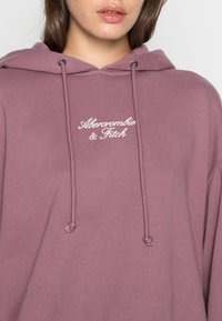 Sudadera morada con cordón, con "Abercrombie & Fitch" bordado en blanco. Textura suave, ajuste relajado y capucha.