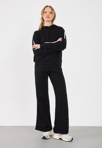 Czarna bluza z kapturem z białymi paskami na rękawach, w zestawie z pasującymi czarnymi spodniami o szerokich nogawkach. Model nosi szare buty sportowe.