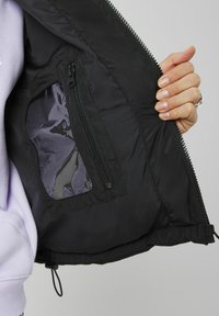 Intérieur de veste noire avec une poche zippée et une superposition transparente. Texture lisse, ourlet élastique en bas, et détails de fermeture éclair visibles.