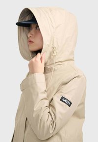 Beige isolierte Jacke mit großer Kapuze, verstellbarem Kordelzug und schwarzem Logo-Patch am Ärmel. Glatte Textur mit modernem Design.