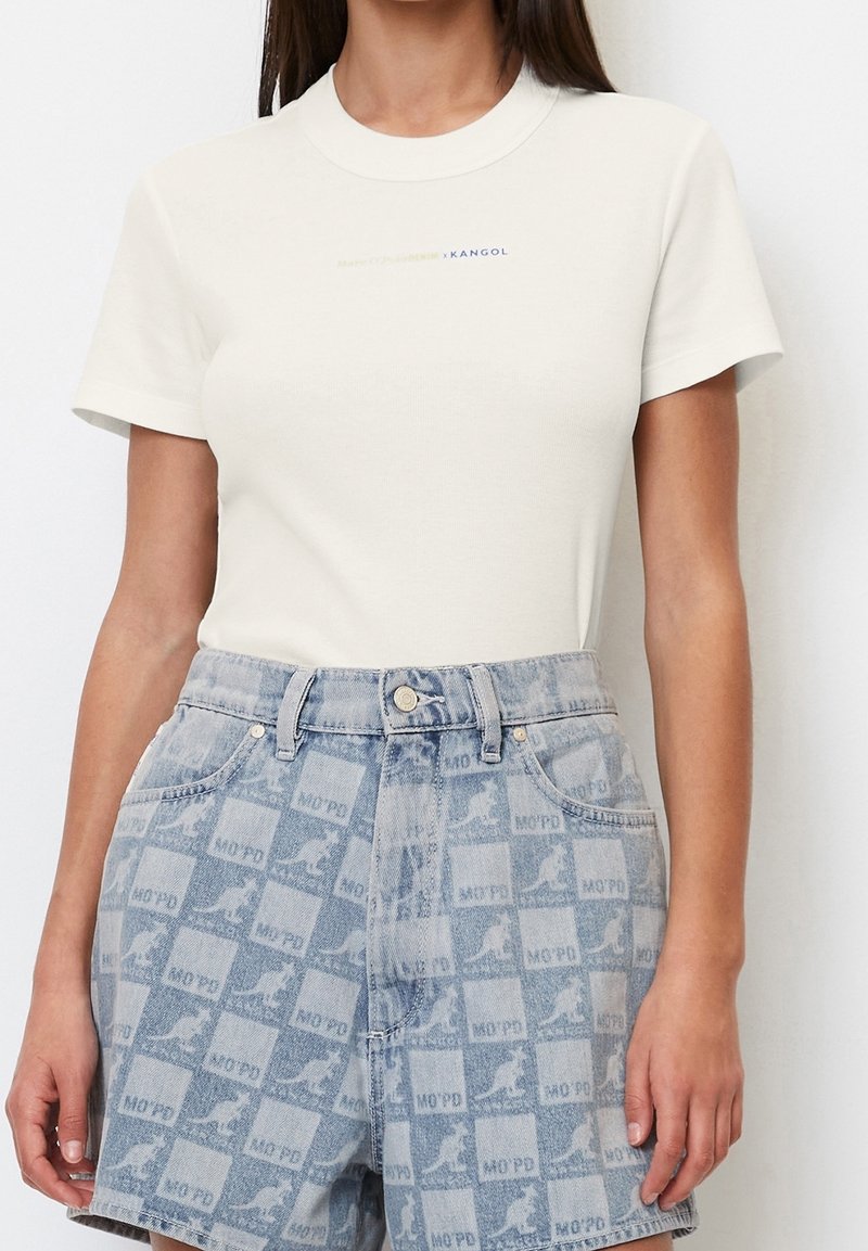 Kvinde iført hvid kortærmet skjorte stoppet ned i høj talje blå denimshorts med kenguru-logo og ternet mønster.