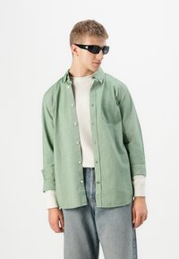 Joven con gafas de sol negras, camisa de botones verde claro sobre una camiseta de manga larga beige, y jeans azul claro, mirando hacia su izquierda.