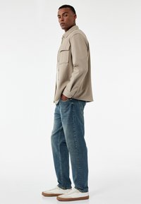 Camisa beige de botones con dos bolsillos en el pecho, combinada con jeans de mezclilla azul y zapatillas blancas con suelas marrones. Textura de tela suave.