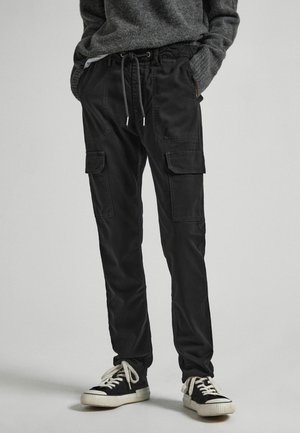 Person trägt schwarze Cargohosen mit Kordelzug in der Taille und vorderen Klappentaschen, kombiniert mit schwarzen und weißen Sneakers und einem grauen Pullover.