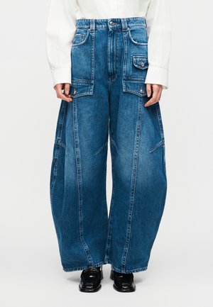 Άτομο που φοράει μπλε καμπάνες τζιν παντελόνι με μεγάλες τσέπες με καπάκι, λευκό μακρυμάνικο πουκάμισο και μαύρα παπούτσια, στέκεται όρθιο.