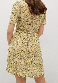 Ein Blumen Kleid in Gelb- und Grüntönen, mit kurzen Ärmeln, tailliertem Bund und einem fließenden Rock aus einem strukturierten, leichten Stoff.