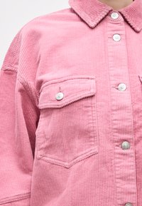 Κοντινή λήψη ενός ροζ μπουφάν κορδούρας με ασημένια κουμπιά πρόσκρουσης και τσέπη στο στήθος με καπάκι.