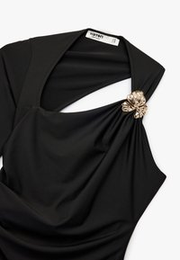 Sort kjole lavet af glat stof, med et draperet design, udskåret skulder og en gylden blomsterdetalje ved stroppen.