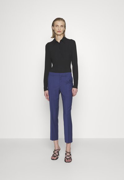 Cămașă polo neagră cu mâneci lungi și textură ribbed, cu guler, purtată cu pantaloni albaștri ajustați până la gleznă și sandale negre cu bretele.