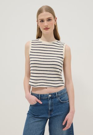 Giovane donna con lunghi capelli biondi che indossa un top crema senza maniche con strisce nere orizzontali e jeans blu a vita alta, in piedi.