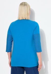 Blaues Baumwolloberteil mit dreiviertellangen gestreiften Ärmeln. Verfügt über einen runden Ausschnitt und eine glatte Textur, die bis zur Hüfte reicht.