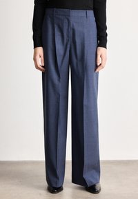 Persona valkā augsta vidukļa, platām kāju zilām kleitas biksēm ar priekšējām krokām, kopā ar melniem, smailiem apaviem un melnu ar garām piedurknēm topu.