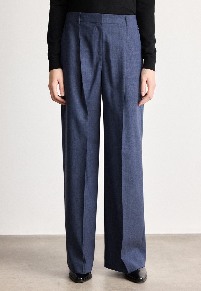 Persona valkā augsta vidukļa, platām kāju zilām kleitas biksēm ar priekšējām krokām, kopā ar melniem, smailiem apaviem un melnu ar garām piedurknēm topu.