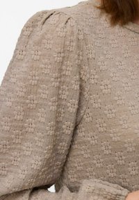 Bežasta teksturirana bluza s nabranim rukavima, koja sadrži cvjetni uzorak i tkaninu od pletene pređe. Šivani detalji dodaju dimenzionalnost dizajnu.