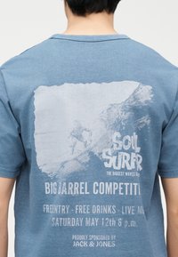 Πίσω μέρος μπλε T-shirt με ξεθωριασμένη λευκή εκτύπωση ενός σερφέρ που καβαλάει ένα κύμα, κείμενο για τον διαγωνισμό "Soil Surfer", ελεύθερη είσοδος, ποτά, ζωντανή μουσική, εκδήλωση στις 12 Μαΐου.