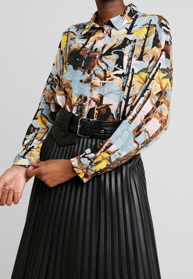 Femme portant une chemise à boutons avec un imprimé abstrait multicolore rentrée dans une jupe plissée en cuir noir avec une large ceinture.