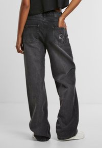 Zwarte denim jeans met wijde pijpen, vijf zakken, contrasterende stiksels en een verhoogd logo detail op de achterzak. Vlotte textuur.
