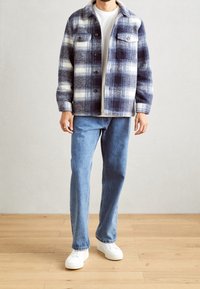 Giacca in flanella a quadri blu navy e bianca, con chiusura a bottoni, due tasche sul petto, abbinata a jeans azzurri chiari e sneakers bianche.