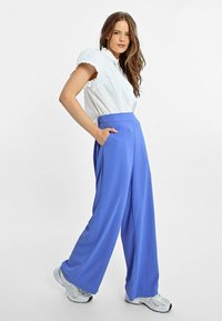Pantaloni ampi azzurri con vita alta e tasche laterali, abbinati a una camicia bianca a maniche corte con dettagli a volant sulle spalle.
