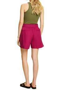 Pantalones cortos fucsias con cintura elástica, dos bolsillos traseros y corte relajado, combinados con una camiseta sin mangas verde y sandalias negras.