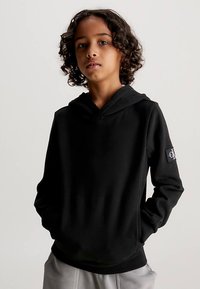 Felpa nera con cappuccio, tasca frontale e orlo arrotondato. Presenta una toppa con logo sulla manica sinistra e un tessuto morbido e testurizzato.