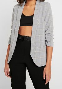 Veste à carreaux grise à manches longues avec poignets retroussés, col cranté et poches latérales, portée sur un haut noir et un pantalon noir.