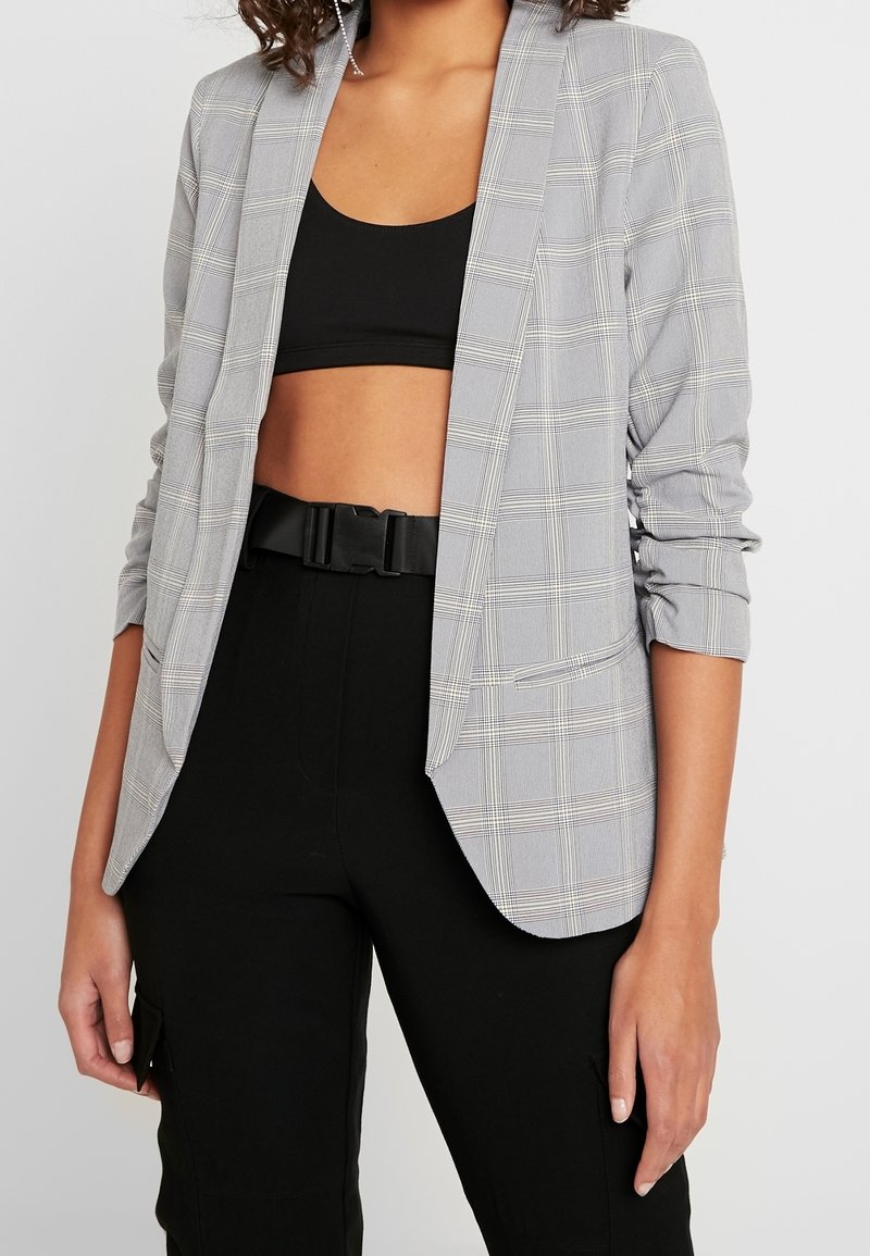 Veste à carreaux grise à manches longues avec poignets retroussés, col cranté et poches latérales, portée sur un haut noir et un pantalon noir.