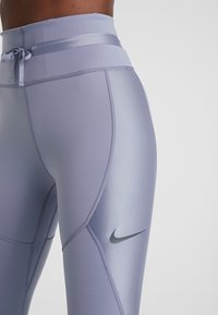 Světle fialové sportovní legíny s vysokým pasem, stahovací šňůrkou, hladkým povrchem a viditelným šedým logem Nike na boku.