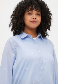 Gaiši zila pogāja krekls ar apkakli, garām piedurknēm un baltiem izšūtiem loku rakstiem visā krekla virsmā. Gluda auduma tekstūra.