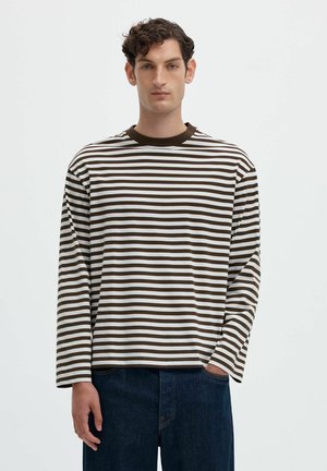Giovane uomo con capelli ricci che indossa una maglietta a maniche lunghe a righe orizzontali marroni e bianche e jeans blu scuro, in piedi davanti a uno sfondo bianco.