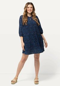 Tumma sininen mekko, jossa on värikkäitä pilkkuja, kerroksellinen muotoilu, kolme neljännespituista puffihihnaa ja avainaukko kaula-aukko. Yhdistetty vaaleanruskeisiin espadrilloihin.